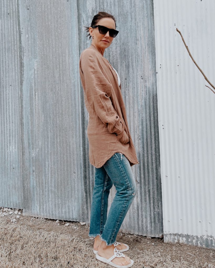 woman looking over her sholder standing by tin shed wearing brown button up and jeans